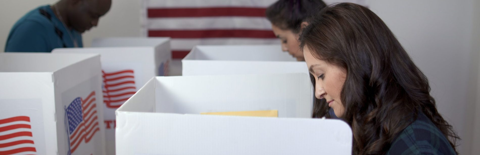 Side view MS of three people, two women and one man, Caucasian, Hispanic and African American, voting in booths at polling station. Large US flag on wall behind